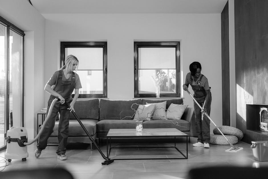Two professional cleaning staff members stand back-to-back against a plain white wall, dressed in beige cleaning uniforms. The woman on the left, with dark hair tied back, holds a red vacuum cleaner. 