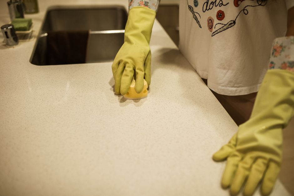 Close-up view of a person wearing yellow rubber gloves holding a blue kitchen sponge in one hand and a clear plastic spray bottle with a red nozzle in the other, prepared for surface cleaning. The bac